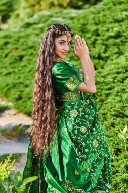 Yeşil sarili uzun saçlı, sırıtan Hintli kadın parkta bitkilerin yanında poz veriyor.