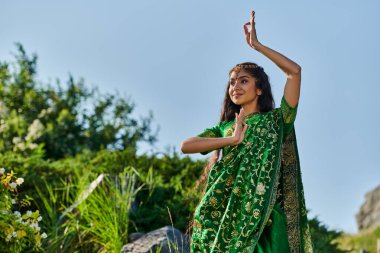 Arka planda gökyüzü ile parkta dikilirken şık yeşil sari giymiş gülümseyen Hintli kadın.