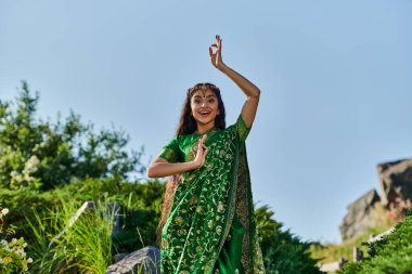 heyecan verici genç Hintli kadın şık yeşil sari giymiş yaz parkında arka planda poz veriyor.