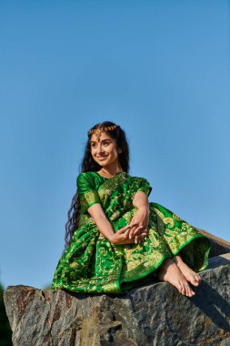 Kaygısız genç Hintli kadın yeşil sari içinde taşın üzerinde oturuyor ve gökyüzünü arka planda seyrediyor.