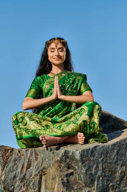 Gülümseyen Hintli kadın sari içinde meditasyon yaparken arka planda mavi gökyüzü olan bir taşın üzerinde oturuyor.