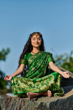 Çıplak ayaklı gülümseyen Hintli kadın sari içinde taşın üzerinde meditasyon yapıyor. Arka planda mavi gökyüzü.