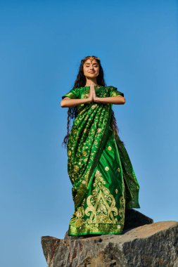 Şık giyinmiş genç Hintli kadın taşın üzerinde meditasyon yapıyor. Arka planda mavi gökyüzü.