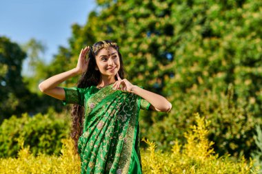 trendy young indian woman in traditional outfit and bindi posing near plants in park in summer clipart