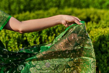 cropped view of young woman touching modern green sari with pattern near plants in park clipart