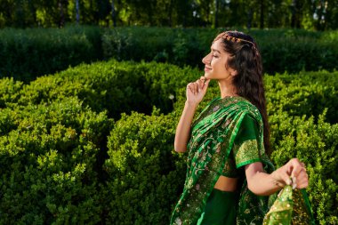 Modern sari içinde yeşil bitkilerin yanında duran genç ve şık Hintli kadının yan görüntüsü.