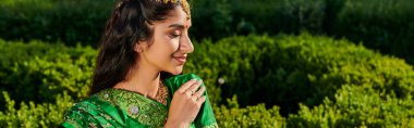 side view of elegant young indian woman in green sari standing near blurred plants in park, banner clipart