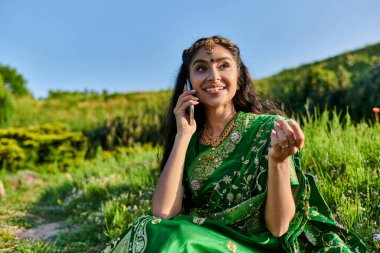Moda elbisesi içinde neşeli, genç Hintli bir kadın yazın çimenlerde otururken akıllı telefonuyla konuşuyor.