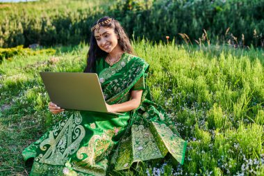 stylish and smiling young indian woman in sari using laptop while sitting on meadow in summer clipart