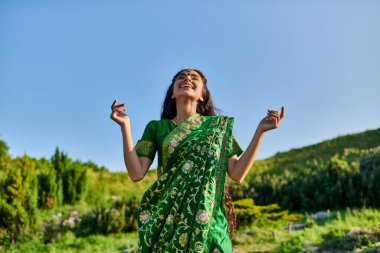 carefree indian woman in sari and matha patti while standing with summer landscape on background clipart