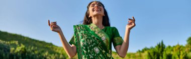 happy young indian woman in green sari standing with summer landscape on background, banner clipart