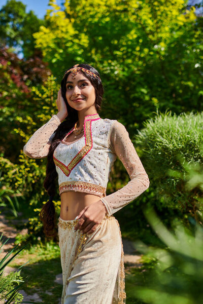 young and pleased woman in elegant traditional clothes looking away in summer park