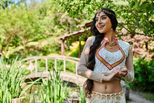 Şaşkın ve mutlu etnik giyimli Hintli kadın yeşil yaz parkında gözlerini kaçırıyor.