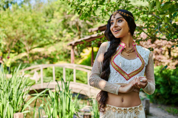 изумленная и счастливая индийская женщина в элегантной этнической одежде смотрит вдаль в зеленом летнем парке