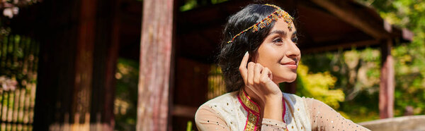 brunette, smiling and dreamy indian woman in traditional clothes looking away in summer park