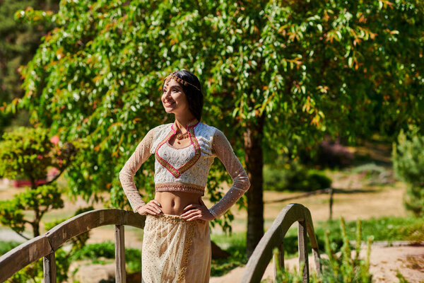 joyful indian woman in traditional wear standing with hands on hips on wooden bridge in green park