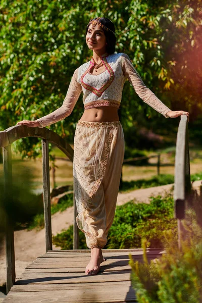 日当たりの良い公園の木の橋の上に立っているエレガントな伝統的な服のブルネットインドの女性 — ストック写真