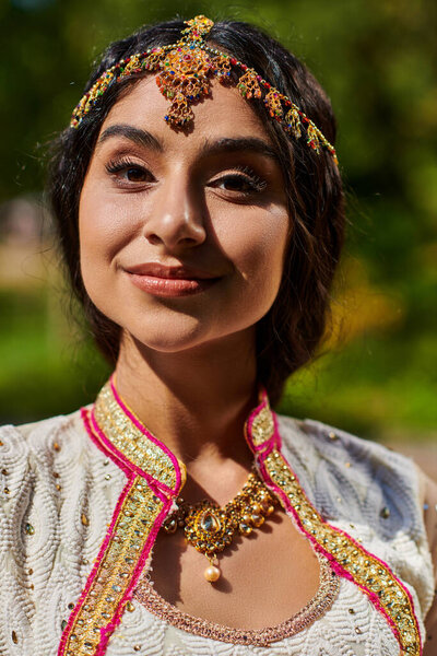 portrait of young brunette indian woman in authentic attire smiling at camera on summer day in park
