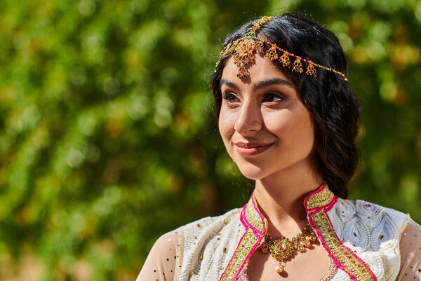 portrait of pleased, brunette indian woman in ethnic wear looking away and smiling in summer park