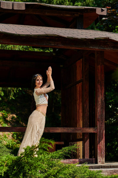 smiling indian woman in traditional attire posing with praying hands near wooden alcove in park