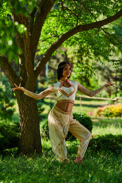 graceful indian woman in vibrant ethnic attire dancing under tree in summer park, happiness