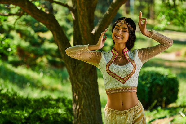 young and happy indian woman in authentic wear dancing in park on sunny day, summer happiness