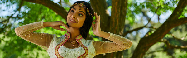 cheerful indian woman in vibrant authentic attire posing and looking away in park, banner