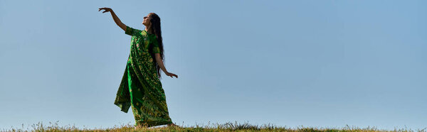 young indian woman in traditional sari in green field under blue and clear sky, summer, banner