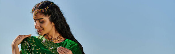 улыбающаяся молодая индийская женщина с Матха Патти касаясь зеленого сари с голубым небом на заднем плане, баннер