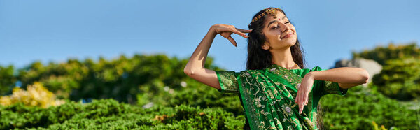 веселая индийская женщина в матха-патти и сари позирует рядом с растениями с голубым небом на заднем плане, баннер