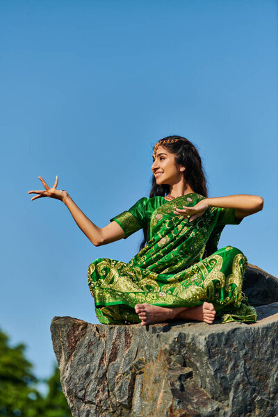 cheerful and stylish indian woman in green sari posing on stone with blue sky on background
