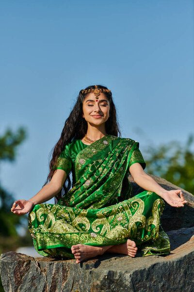 smiling indian barefoot woman in sari meditating on stone with blue sky on background