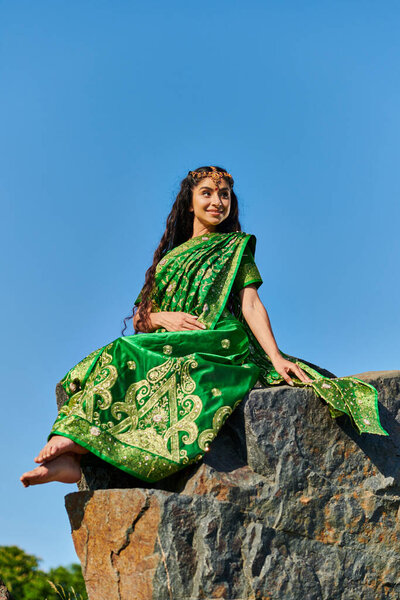 carefree barefoot indian woman in stylish green sari sitting on stone with sky on background