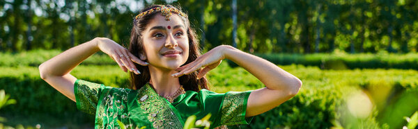 улыбающаяся молодая индийская женщина с бинди и сари позируют рядом с растениями в парке летом, баннер