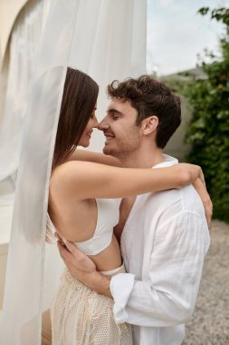 Seksi çift, mutlu adam güzel bir kadını kucaklarken plajdaki beyaz tülün yanında duruyor.