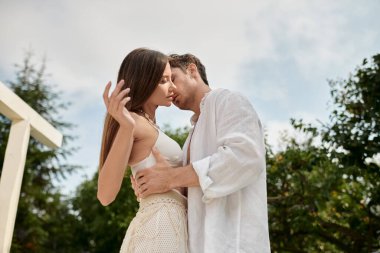 passionate man embracing pretty woman in white beach wear on luxury resort during vacation clipart