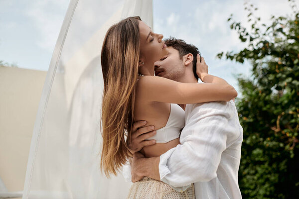 passionate couple, man kissing neck of woman while standing near white tulle of pavilion on beach