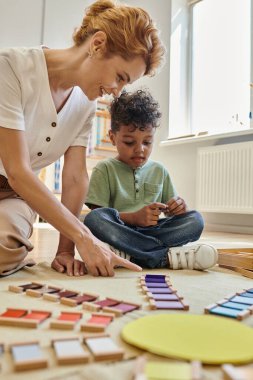 Montessori okulu, mutlu öğretmen Afrika kökenli Amerikalı çocuğun yanındaki tahta renkli blokları gösteriyor.