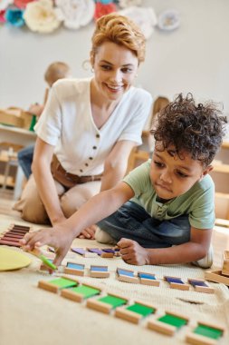 Montessori materyali, zeki Afrikalı Amerikalı çocuk gururlu öğretmenin yanında eğitici renk oyunu oynuyor.