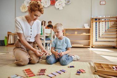 Montessori okulu, bayan öğretmen sarışın çocuğun yanında oturuyor ve tahta oyuncaklar gösteriyor, eğitici bir oyun.