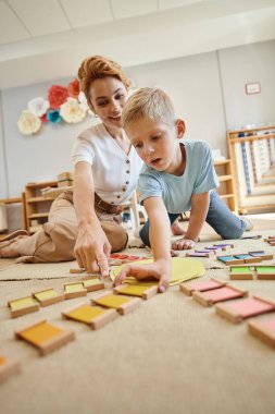 Montessori okulu, sarışın çocuk kadın öğretmenin yanında eğitim oyunu oynuyor, hareket, renk eşleşmesi.