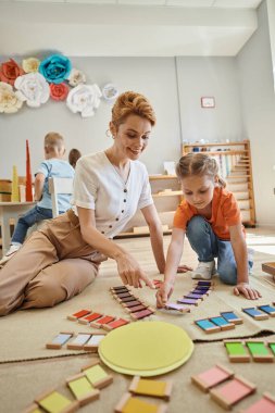 Montessori okulu, renkli oyun oynayan tatlı bir kız mutlu bayan öğretmenin yanında, yerde oturuyor.