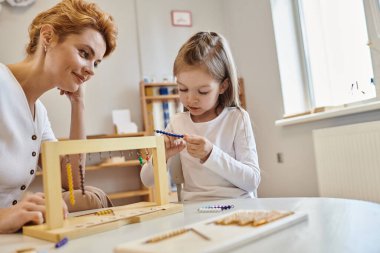 Montessori konsepti, öğretmenlerin yanında renkli boncuk merdivenlerle oynayan kız, ahşap tezgah, eğitici