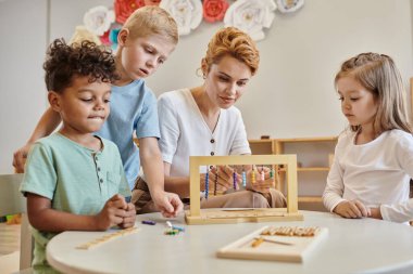 Montessori okulu, ırklar arası çocukları gözlemleyen kadın öğretmen, eğitim oyunu oynayan, çeşitli çocuklar.