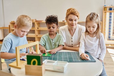 Montessori okulu konsepti, çok kültürlü çocuklar, öğretmen ve erkek çocuklarının yanındaki tahtaya yazı yazan kız.