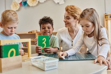 Mutlu Afro-Amerikan çocuk elinde kartonu tutuyor. Yanında çocuklar ve Montessori okulundaki öğretmen var.