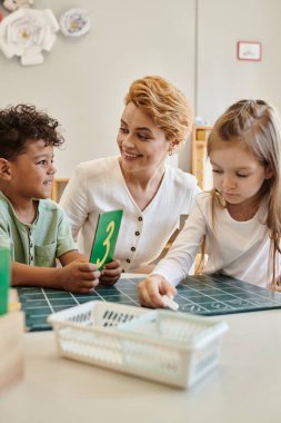Mutlu Afrikalı Amerikalı çocuk Montessori okulundaki kız ve öğretmenin yanında karton tutuyor.