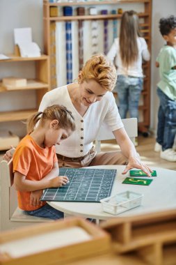 Sayıların yakınındaki tahtaya yazı yazan kız oyun, öğretmen ve çocuklarla oyun oynuyor. Montessori konsepti.