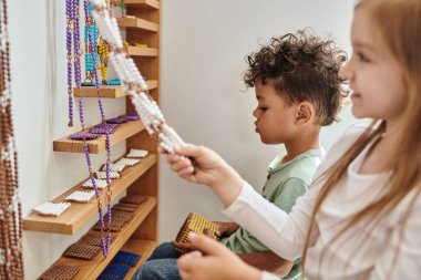 cheerful girl pulling beads near african american boy, diversity, Montessori school concept clipart