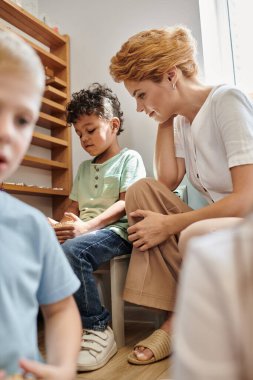 Sarışın öğretmen küçük Afrikalı Amerikalı çocuğu gözlemliyor, Montessori okulu, oyun oynayarak öğreniyor.